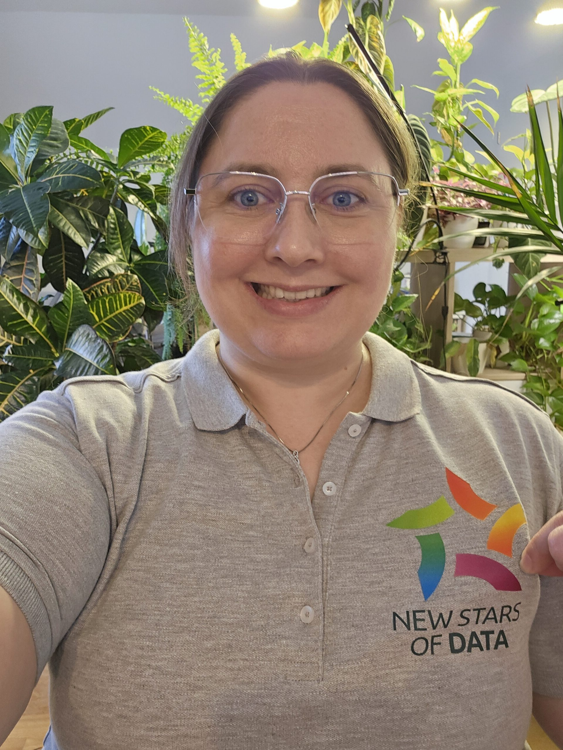 Karin Szilágyi lächelt vor Zimmerpflanzen im Hintergrund und trägt ein graues New Stars of Data Poloshirt nach ihrer erfolgreichen Konferenz-Session.